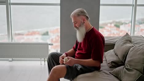 Ein-älterer-Mann-Mit-Grauem-Haar-Und-Buschigem-Bart-Verbindet-Sein-Knie,-Während-Er-Auf-Dem-Sofa-In-Einem-Modernen-Apartment-Mit-Blick-Aufs-Meer-Sitzt