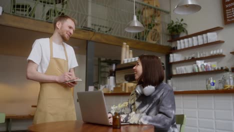 Un-Camarero-Alegre-Con-Un-Delantal-Marrón-Claro-Toma-El-Pedido-De-Una-Chica-Morena-Que-Está-Trabajando-Frente-A-Una-Computadora-Portátil-En-Una-Mesa-De-Un-Café.