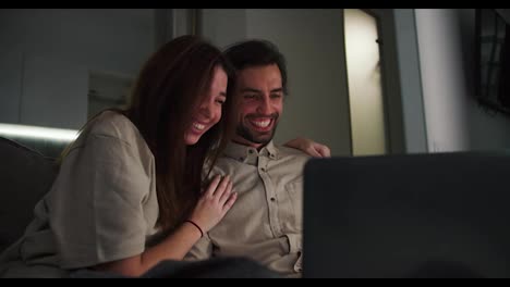 A-happy-brunette-girl-in-a-beige-T-shirt-hugs-her-cheerful-brunette-boyfriend-with-stubble-they-watch-a-comedy-together-on-a-gray-laptop-in-the-evening-in-a-modern-studio-apartment