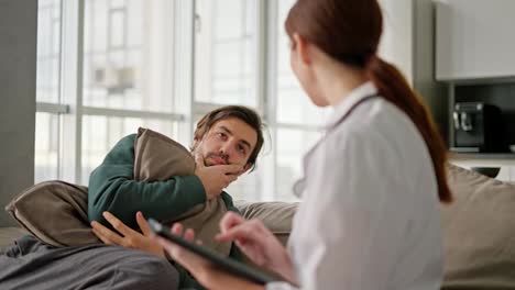 Un-Hombre-Moreno-Con-Barba-Incipiente-Y-Una-Chaqueta-Verde-Le-Cuenta-A-Una-Doctora-Morena-Con-Una-Bata-Médica-Sobre-Sus-Problemas-Mientras-Está-Sentado-En-El-Sofá-Y-Se-Abraza-En-Un-Apartamento-Moderno-Durante-Un-Examen-Domiciliario.