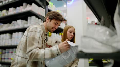 A-happy-couple-a-brunette-guy-and-a-blonde-girl-in-checkered-shirts-together-with-their-white-dog-inspect-items-in-a-pet-store-and-choose-the-one-they-need