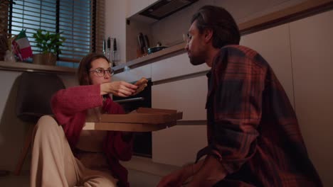 A-brunette-girl-with-glasses-in-a-red-sweater-feeds-her-brunette-boyfriend-with-stubble-pizza-from-delivery-Sitting-on-the-floor-in-a-modern-apartment-in-the-kitchen-in-the-evening