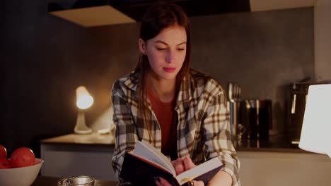 Happy-brunette-girl-in-a-plaid-shirt-and-an-orange-T-shirt-stands-in-the-kitchen-and-reads-a-book-during-her-evening-relaxation-in-a-modern-apartment