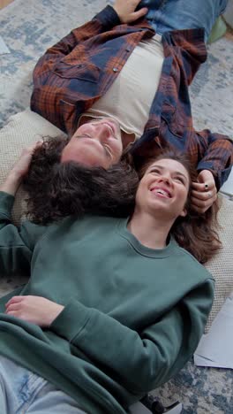 Vertical-video-of-a-happy-brunette-girl-in-a-green-jacket-lies-head-to-head-with-a-guy-in-a-checkered-shirt-on-the-floor-and-smiles