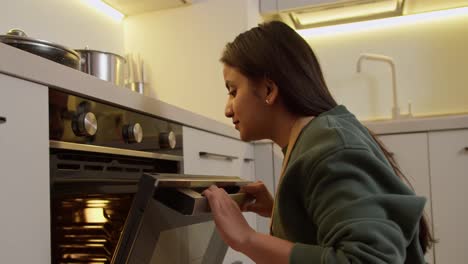 Happy-and-confident-housewife-brunette-girl-with-Brown-skin-color-in-a-green-jacket-and-apron-checks-the-readiness-of-food-in-the-oven-during-her-cooking-at-home-in-a-modern-apartment