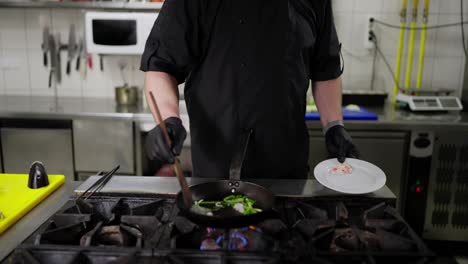 A-man-in-a-black-professional-chef-uniform-adds-ingredients-to-a-frying-pan-and-fries-the-ingredients-until-done-on-the-stove-in-the-kitchen-in-a-restaurant