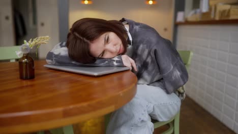 A-tired-brunette-girl-in-a-gray-sweater-a-cafe-visitor-lies-on-a-laptop-at-the-table-and-sleeps-in-a-cafe