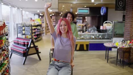 Retrato-De-Una-Niña-Feliz-Con-Cabello-Rosado-Siendo-Empujada-En-Un-Carrito-Por-Su-Novio-Mientras-Juega-Y-Se-Divierte-En-El-Supermercado.