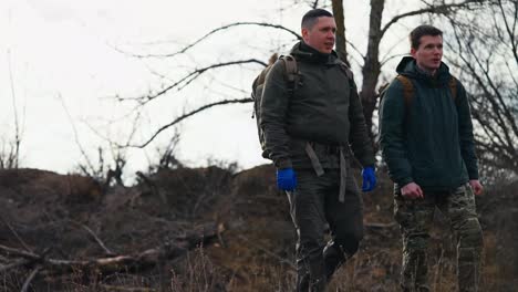 Vista-Lateral-De-Dos-Jóvenes-Confiados-Con-Uniformes-Militares-Verde-Oscuro-Y-Guantes-Médicos-Azules-Caminando-Por-El-Campo-De-Entrenamiento-Militar-Y-Prácticas-De-Combate-Y-Comunicándose-Con-Mochilas-En-Sus-Espaldas.