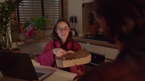 Confident-young-brunette-girl-with-glasses-in-red-wireless-headphones-and-a-sweater-holds-a-box-along-with-pizza-in-her-hands-and-makes-a-snack-during-her-foreign-language-lesson-with-a-teacher-in-the-kitchen-in-a-modern-apartment