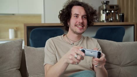 Happy-brunette-guy-with-curly-hair-and-mustache-in-a-gray-T-shirt-plays-on-a-white-joystick-and-sits-on-the-sofa-during-his-time-on-the-weekend