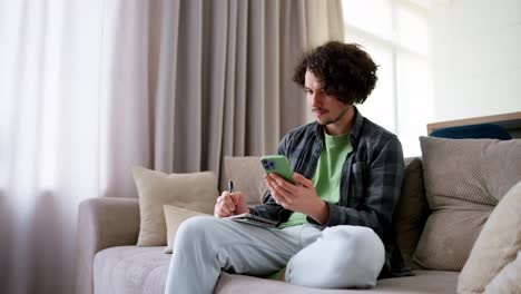 A-confident-brunette-guy-with-curly-hair-in-a-gray-checkered-shirt-takes-notes-and-notes-that-he-watches-a-video-on-a-green-smartphone-at-home-on-the-sofa-in-a-modern-apartment