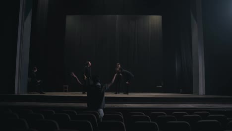 Rear-view-of-a-young-male-production-director-standing-in-the-hall-and-showing-how-to-move-the-actors-and-what-to-do-on-stage-during-a-rehearsal-in-an-empty-theater-with-black-curtains