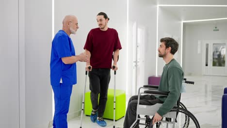 Un-Hombre-Seguro-De-Sí-Mismo-Con-Barba-Gris,-Un-Médico-Con-Uniforme-Azul-Se-Comunica-Con-Hombres-Después-De-Sus-Lesiones-En-Una-Clínica-Moderna-Con-Muletas-Y-Sillas-De-Ruedas-Para-Discapacitados