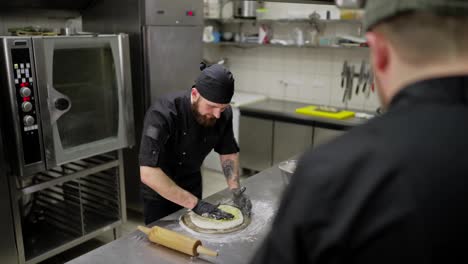 Ein-Selbstbewusster-Männlicher-Pizzaiola-In-Einer-Schwarzen-Uniform-Gibt-In-Der-Küche-Eines-Restaurants-Soße-Auf-Ein-Stück-Pizzateig.-Ein-Professioneller-Männlicher-Koch-In-Einer-Schwarzen-Uniform-Bereitet-Eine-Pizza-Auf-Werkstückhöhe-Zu-Und-Verteilt-Die-Soße-Auf-Der-Oberfläche-Des-Teigs