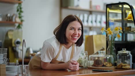 Portrait-of-a-happy-brunette-girl-in-a-white-T-shirt-and-light-brown-apron-who-looks-out-and-leans-on-the-counter-in-a-cafe