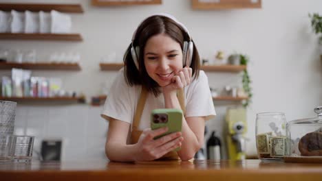 Zoom-in-happy-girl-waiter-in-a-white-t-shirt-listens-to-music-and-watches-a-video-in-white-wireless-headphones-behind-the-counter-in-a-cafe