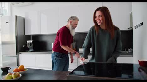 A-man-with-gray-hair-with-a-lush-gray-beard-in-a-red-shirt-together-with-his-adult-brunette-daughter-in-a-green-sweater-is-cleaning-in-a-modern-kitchen