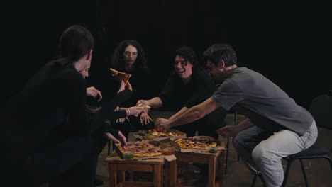 A-group-of-happy-and-confident-actors-in-black-uniforms-gathered-around-pizza-having-lunch-and-chatting-during-a-rehearsal-break-on-the-theater-stage.-A-group-of-happy-actors-gathered-around-the-ordered-pizza-sit-have-lunch-and-chat-about-the-next-scene-and-role-in-the-theater