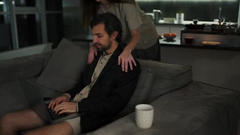 A-happy-brunette-girl-in-a-beige-T-shirt-approaches-her-brunette-boyfriend-with-stubble-in-a-black-jacket-and-beige-shorts-and-asks-how-his-work-is-going-remotely-in-a-modern-apartment-in-the-evening