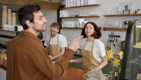 A-happy-brunette-girl-waiter-at-the-checkout-in-a-yellow-apron-takes-an-order-and-gives-it-to-a-blond-guy-nearby-while-serving-a-client-in-a-cafe