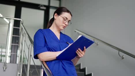 Confident-brunette-girl-in-round-glasses-doctor-in-a-blue-uniform-takes-notes-on-a-blue-tablet-while-standing-on-the-stairs-in-a-modern-clinic