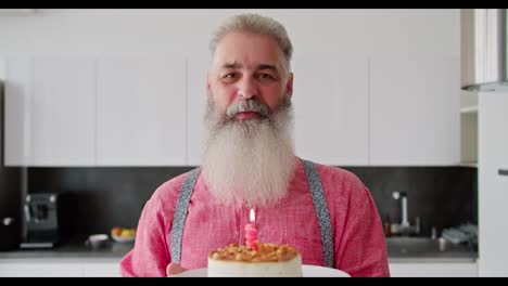 Portrait-of-a-happy-elderly-man-with-gray-hair-with-a-lush-beard-in-a-pink-shirt-who-holds-a-cake-with-a-burning-candle-in-front-of-him-and-blows-it-out-in-honor-of-his-birthday-in-a-modern-kitchen