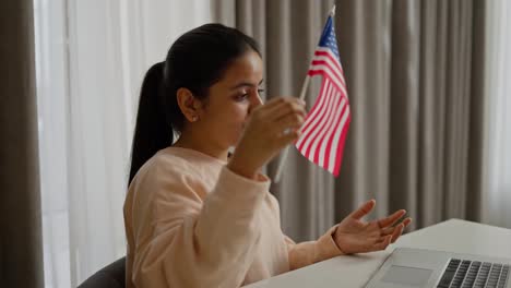 Side-view-of-a-happy-brunette-girl-in-a-beige-sweater-studying-foreign-languages-online-in-front-of-a-computer-at-home-and-showing-the-flag-of-the-United-States-of-America-to-the-webcam-in-a-modern-apartment-during-the-day