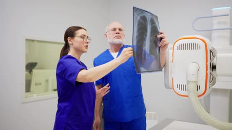 A-confident-brunette-girl-with-glasses-in-a-blue-uniform-communicates-together-and-consults-with-a-confident-man-doctor-with-gray-hair-while-summing-up-the-results-of-fluorography-and-confirming-the-diagnosis-in-a-modern-clinic