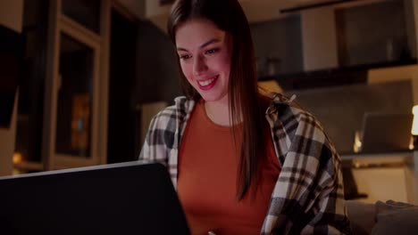 Una-Chica-Morena-Alegre-Y-Segura-De-Sí-Misma-Con-Una-Camisa-A-Cuadros-Y-Una-Camiseta-Naranja-Se-Sienta-En-Un-Sofá-Gris-Y-Mira-Videos-Divertidos-Usando-Una-Computadora-Portátil-En-Un-Moderno-Apartamento-Tipo-Estudio-Por-La-Noche.