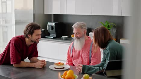 Un-Hombre-Moreno-Feliz-Con-Barba-Incipiente-Con-Una-Camiseta-Roja-Junto-Con-Su-Hermana-Morena-Con-Un-Suéter-Verde-Felicitan-A-Su-Anciano-Padre-Con-Una-Barba-Exuberante-Con-Una-Camisa-Rosa-En-Su-Cumpleaños-Y-Juntos-Apagan-Una-Vela-En-Un-Pequeño-Pastel-Mientras-Están-Sentados-En-Una-Cocina-Moderna-En-Un-Departamento.