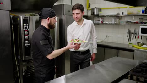 A-confident-male-cook-in-a-black-uniform-gives-the-waiter-a-ready-made-salad-before-serving-the-dish-in-the-restaurant-hall.-A-confident-guy-in-a-white-shirt-brings-the-salad-from-the-chef-into-the-restaurant-hall