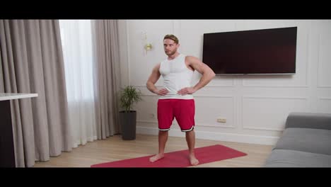 Side-view-of-a-confident-blond-athletic-man-with-stubble-wearing-a-white-T-shirt-and-red-shorts-stands-on-a-red-sports-mat-and-warms-up-during-his-morning-exercises-at-home-in-a-modern-apartment