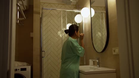 Side-view-of-happy-brunette-Indian-girl-in-Green-pajamas-brushes-her-teeth-while-getting-ready-for-bed-in-the-bathroom-in-a-modern-apartment-in-the-evening