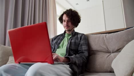 Confident-and-happy-brunette-guy-in-a-checkered-shirt-with-a-mustache-and-curly-hair-sits-on-the-sofa-and-works-on-a-red-laptop-at-home