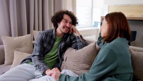 Happy-brunette-guy-with-curly-hair-in-a-gray-checkered-shirt-sits-on-the-sofa-and-communicates-with-a-brunette-girl-while-spending-time-together-in-a-modern-apartment