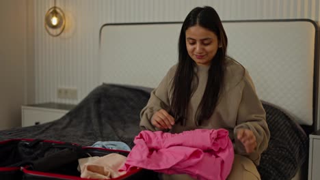 A-happy-and-confident-brunette-girl-with-Brown-skin-in-a-gray-sweater-puts-her-pink-blue-and-beige-things-in-a-black-suitcase-while-preparing-for-the-weekend-and-leaving-travel-in-a-modern-apartment-in-the-bedroom