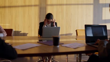 Müdes-Brünettes-Mädchen-Mit-Runder-Brille-Und-Business-Anzug-Sitzt-An-Einem-Holztisch-Im-Büro-Und-Massiert-Ihre-Schläfen-Während-Einer-Pause-In-Der-Angespannten-Atmosphäre-Des-Büros