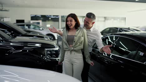 A-happy-middle-aged-man-with-gray-hair-in-a-white-shirt-opens-the-eyes-of-his-happy-brunette-girlfriend-with-his-hands.-I-present-her-with-a-white-car-that-he-bought-for-her-as-a-gift-at-a-car-dealership