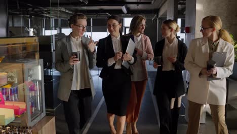 A-group-of-confident-businesswomen-in-business-clothes-walk-along-a-modern-office-during-a-meeting-with-coffee-in-their-hands-and-a-laptop