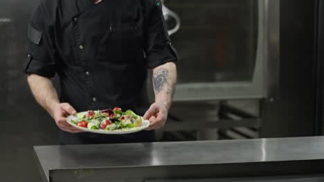 Primer-Plano-De-Un-Chef-Profesional-Con-Uniforme-Negro-Que-Pasa-Un-Plato-De-Ensalada-A-Un-Camarero-Con-Camisa-Blanca-En-La-Cocina-De-Un-Restaurante