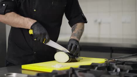 Close-up-a-male-chef-in-a-black-uniform-and-rubber-gloves-carefully-and-skillfully-cuts-an-eggplant-while-preparing-a-dish-on-a-yellow-kitchen-board-in-a-restaurant-kitchen