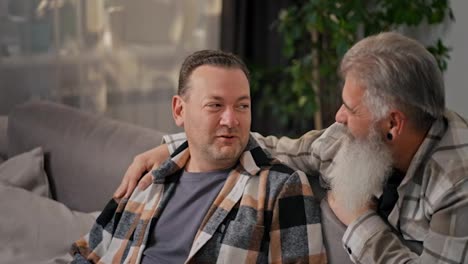 Happy-brunette-man-in-a-checkered-shirt-communicates-with-his-middle-aged-boyfriend-with-gray-hair-and-a-lush-beard-in-a-modern-apartment-on-the-sofa-in-the-evening