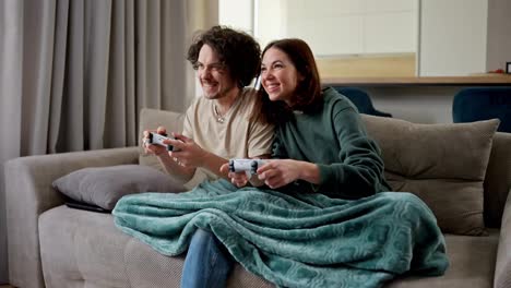 Confident-brunette-guy-with-curly-hair-plays-video-games-using-a-joystick-with-his-girlfriend-in-a-green-jacket-but-the-girl-wins-him-on-the-sofa-in-a-modern-apartment