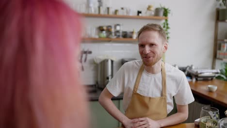 Over-the-shoulder-of-a-happy-blond-guy-in-a-white-T-shirt-and-a-yellow-apron-a-barista-serves-a-client-with-pink-hair-at-the-counter-in-a-cafe