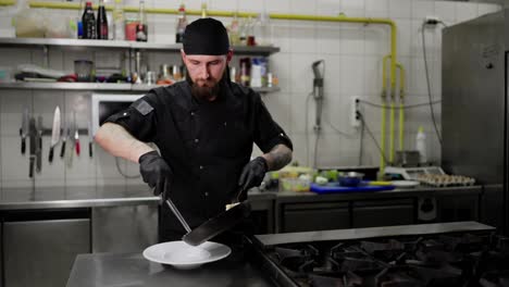 Un-Chef-Profesional-Con-Uniforme-Negro-Coloca-El-Plato-Terminado-En-Un-Plato-Desde-Una-Sartén-Usando-Pinzas-En-La-Cocina-De-Un-Restaurante-Antes-De-Servir-El-Plato.