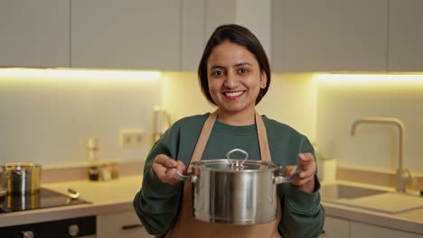 Portrait-of-a-happy-brunette-Indian-girl-in-a-Green-jacket-and-a-beige-apron-holding-a-gray-shiny-pan-in-her-hands-and-looking-at-the-camera-in-a-modern-kitchen