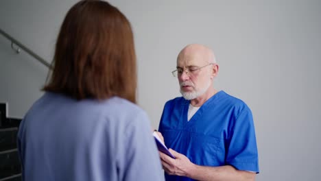 Over-his-shoulder-a-serious-doctor-with-glasses-and-a-gray-beard-in-a-blue-uniform-tells-brunette-girls-about-his-ideas-about-symptoms-and-diagnoses-in-a-modern-clinic