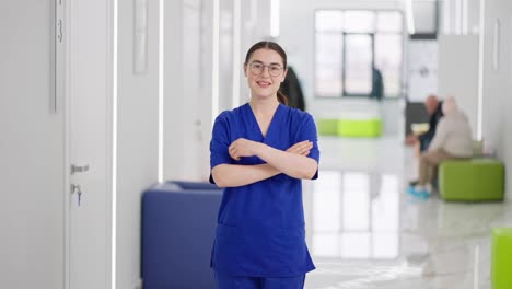 Portrait-of-a-confident-and-happy-brunette-girl-with-glasses-in-a-blue-uniform-the-doctor-crosses-her-arms-over-her-chest-and-poses-in-the-corridor-of-a-modern-light-clinic