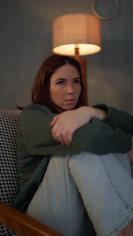 Vertical-video-of-a-sad-confident-brunette-girl-in-a-green-shirt-pressing-her-legs-to-her-body-and-sitting-on-a-chair-near-a-yellow-lamp-at-home